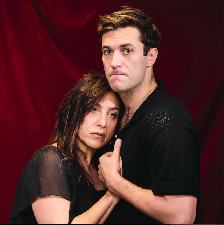 Two performers in black clothing pose together against a deep red curtain backdrop.