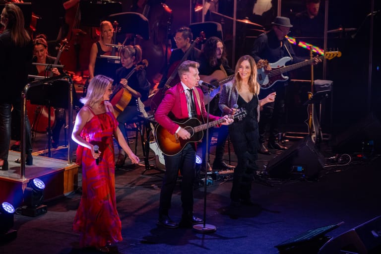 Multiple musicians perform on stage bathed in purple and blue stage lighting, with guitars and drums visible.