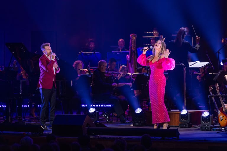Two performers in vibrant jackets on stage with a full band under blue and pink stage lighting.