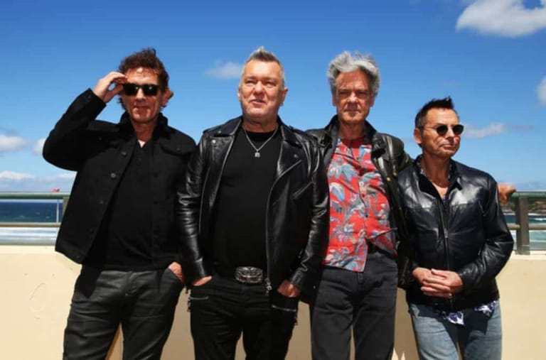 Four musicians in leather jackets stand together on a sunny beach with blue sky behind them.