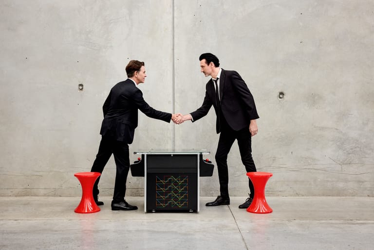 Two men in dark suits shake hands across a black box, standing on red stools against a bare concrete wall.