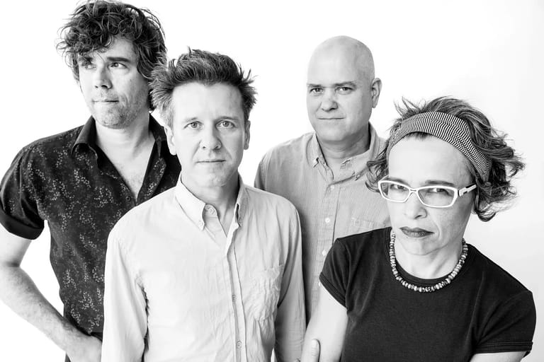 Four musicians posed together against a white background in black and white portrait photography.