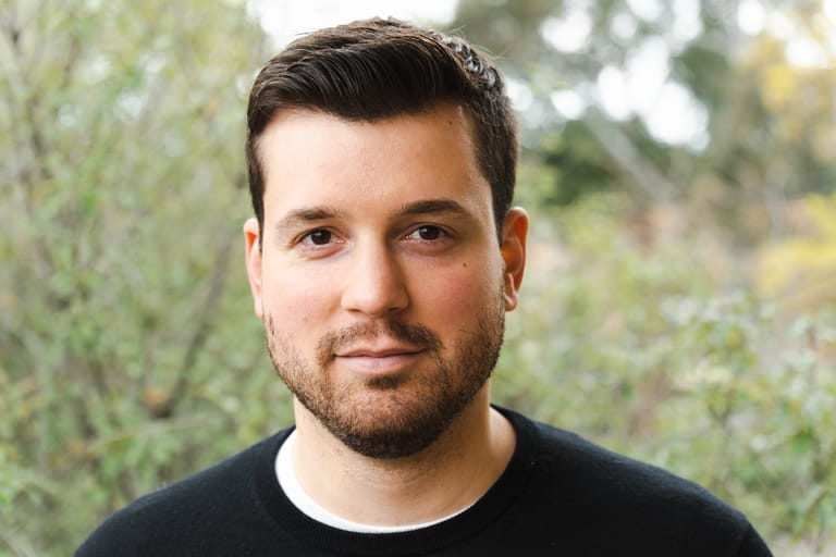 Man with dark hair and beard wearing a black shirt, photographed outdoors against a blurred green leafy background.
