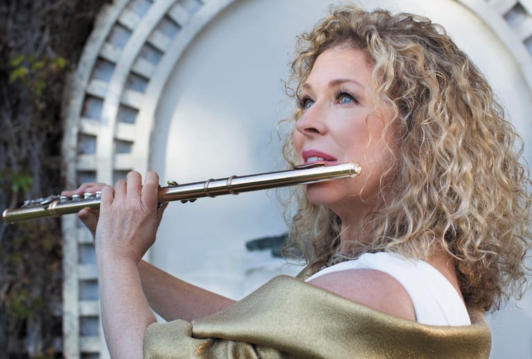 Musician with curly hair plays flute outdoors against a curved architectural backdrop.