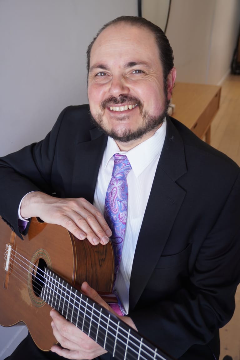 Man in black suit and purple tie holding an acoustic guitar, smiling at the camera.
