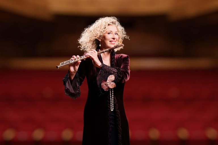 Woman with blonde curly hair singing into microphone on stage, wearing black outfit against red theatre seating background.
