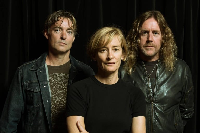 Three musicians pose against a dark backdrop, wearing casual black and leather clothing.