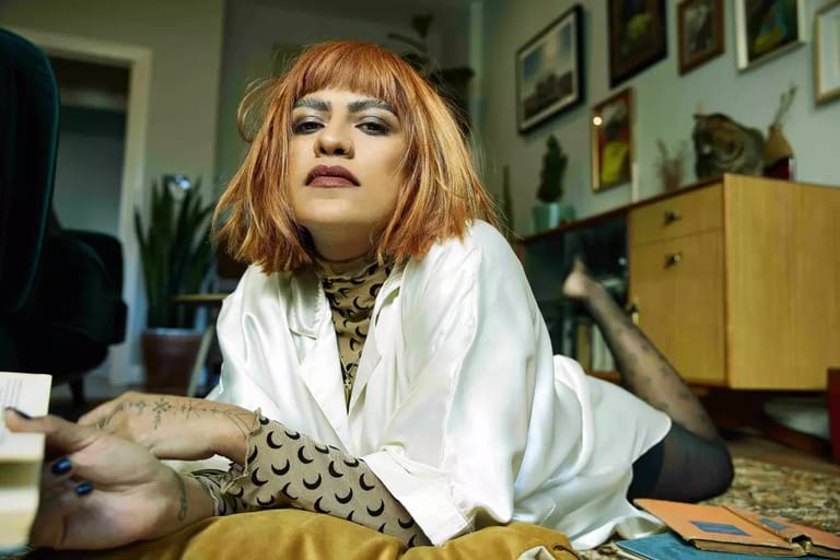 Person with reddish-blonde bob hair and tattoos reclining on a patterned cushion in a vintage-decorated interior space.