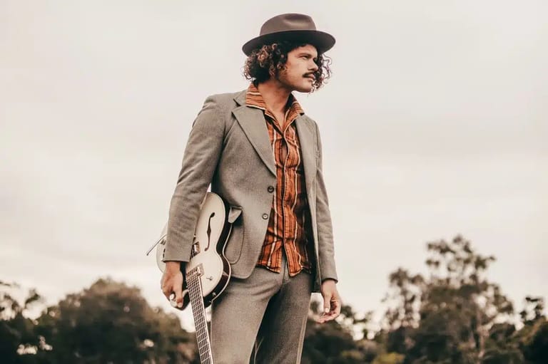 Man in beige suit and fedora hat holding guitar outdoors against tree-lined horizon.