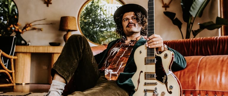 Musician with moustache and hat lounging on a leather sofa while holding a white electric guitar.