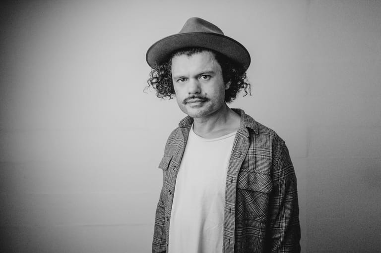 Man with curly hair and moustache wearing a fedora and checked shirt against a grey background.