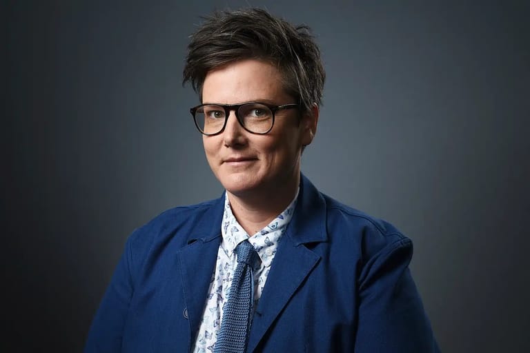 Person wearing glasses and blue blazer against neutral grey background.