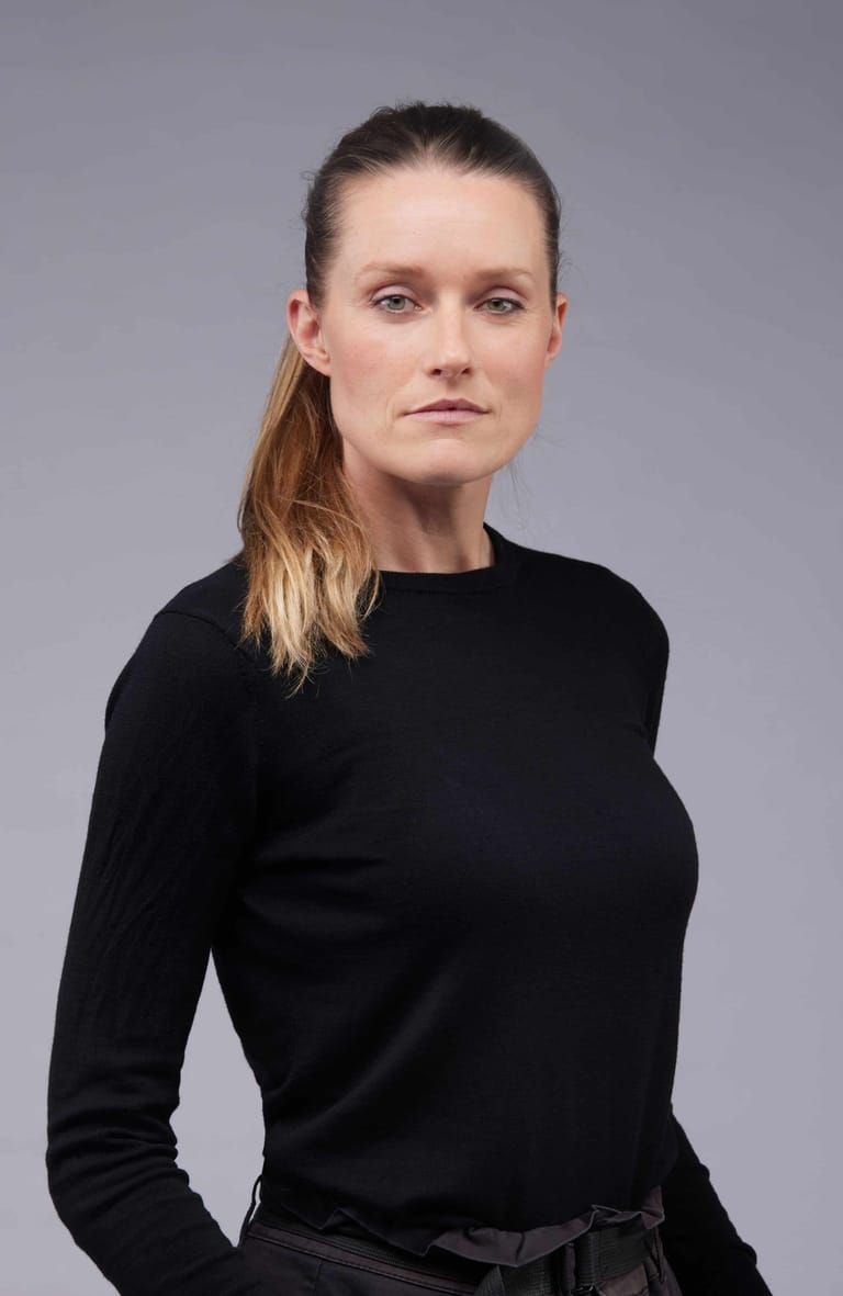 Woman with long auburn hair wearing a black long-sleeved top against a neutral grey background.