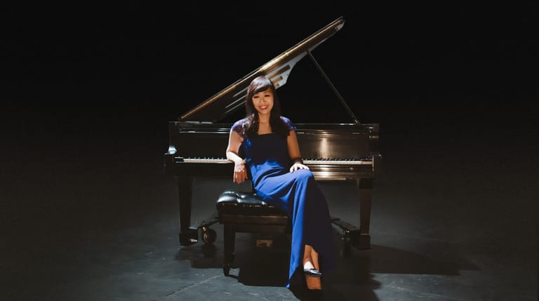 Pianist in a blue dress sits at a grand piano against a dark stage background.