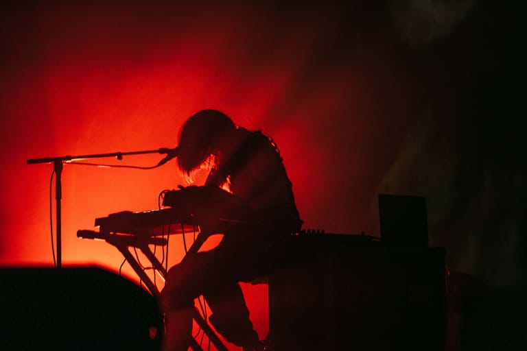 Musician silhouetted against red stage lighting, seated at keyboard with microphone stand nearby.