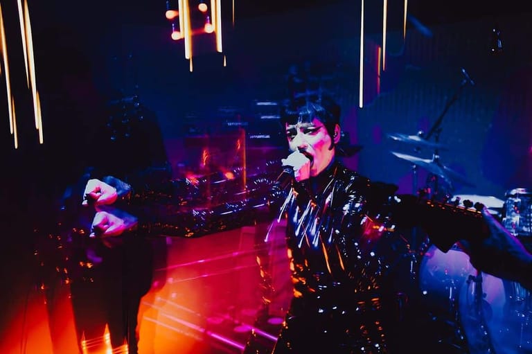Performer in studded leather jacket singing at microphone under red and blue stage lighting with band members visible behind.
