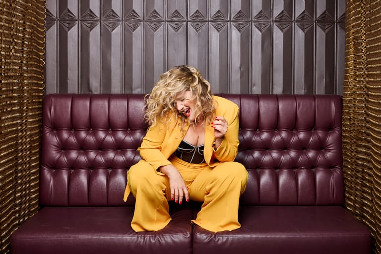 Person in yellow suit crouching on burgundy velvet sofa against brick walls and geometric backdrop.