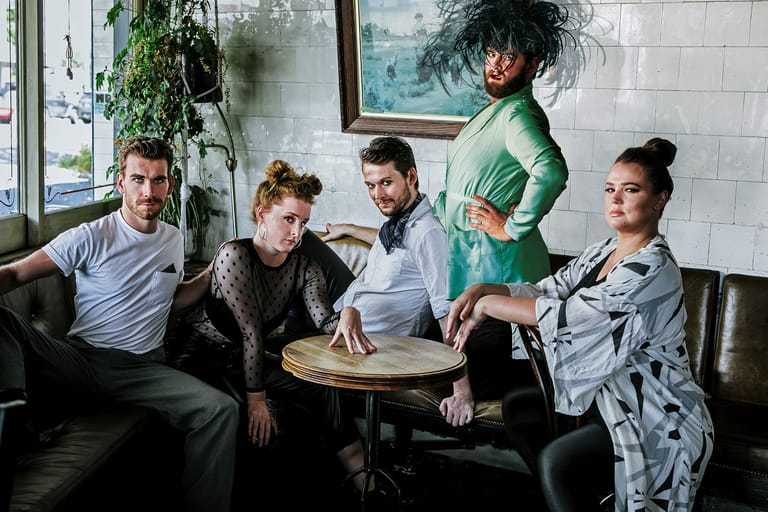 Five people gather around a small table in a casual indoor setting with white tiles and indoor plants visible in the background.