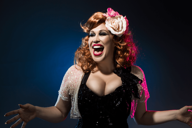 Performer with styled red curled hair and floral headpiece wearing sequined black dress against blue backdrop.
