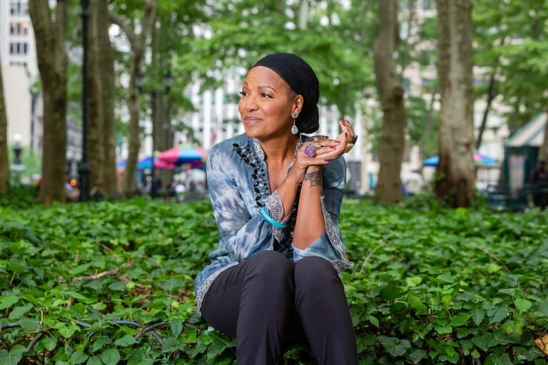 Woman crouching among green foliage in a park, blowing a kiss toward the camera.