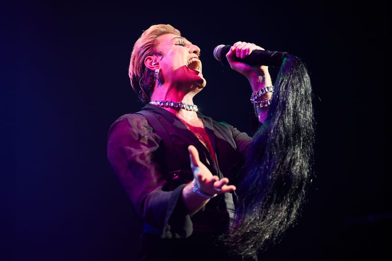 Singer performing at microphone under pink and purple stage lighting, holding long dark hair.