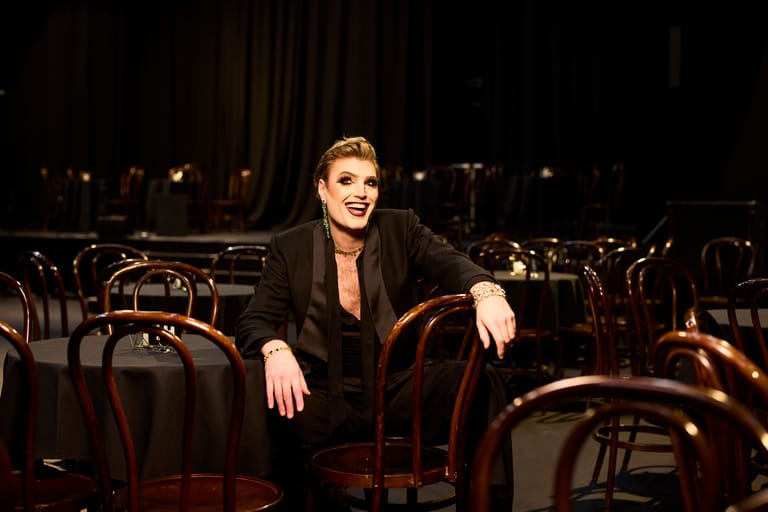Performer in black blazer seated among empty theatre chairs, lit by warm stage lighting.