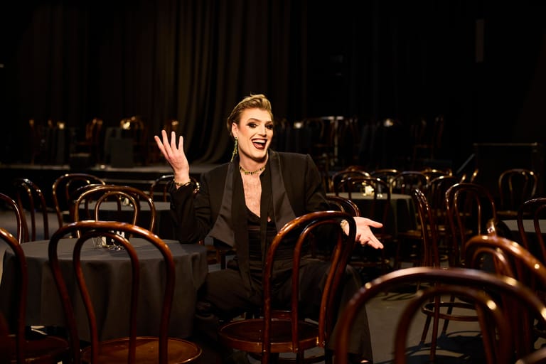 Woman in black blazer gestures expressively while seated among wooden chairs in a darkened theatre space.