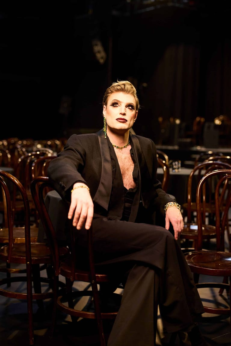 Person in black suit seated amongst wooden chairs on a dimly lit stage.