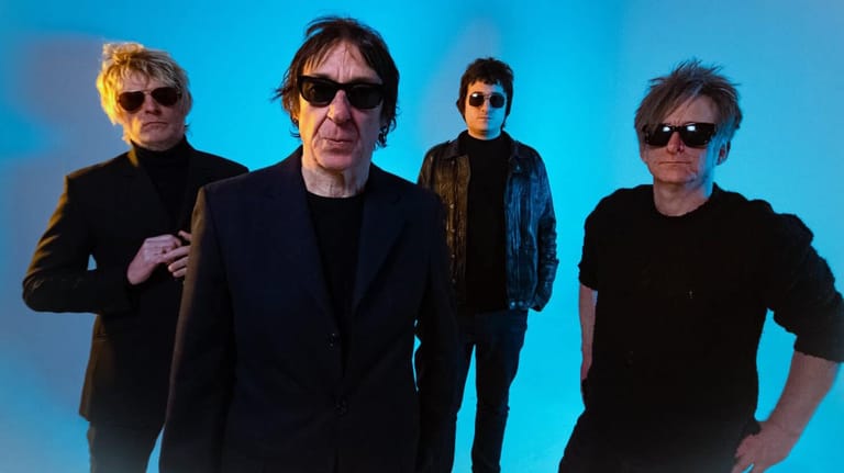 Four musicians in sunglasses and dark clothing posed against a blue background.