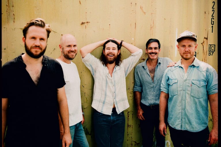 Five male band members stand shoulder-to-shoulder against a weathered yellow wall, smiling at the camera.