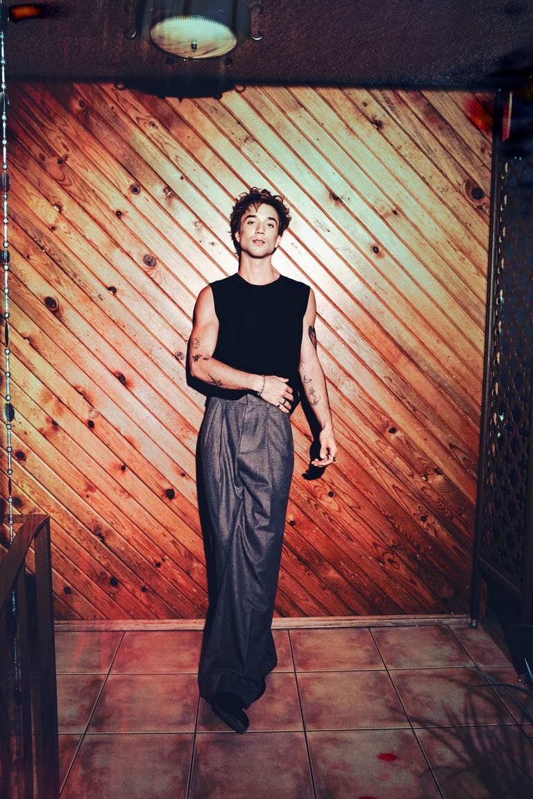 Person in black sleeveless top and grey trousers standing against warm-toned wooden wall.