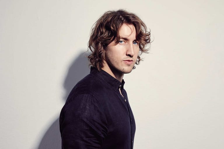 Man with shoulder-length dark wavy hair in dark shirt, photographed in profile against white wall with side lighting.