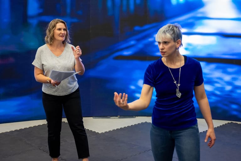 Two women speak on stage under blue lighting, one holding a microphone.