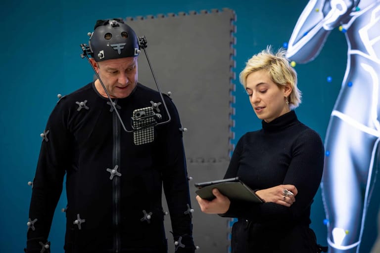 Man in black studded jacket with headgear holds tablet while speaking with blonde woman against blue studio background.
