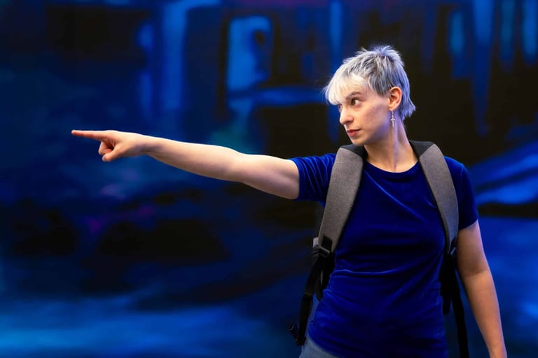 Performer with short grey hair in dark clothing extends arm while pointing across a blue-lit stage.