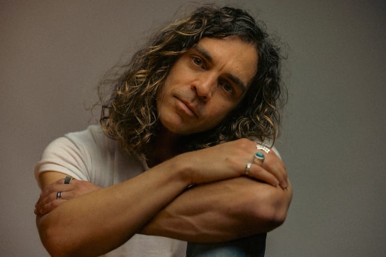 Person with curly dark hair and rings, arms crossed, against a neutral grey background.