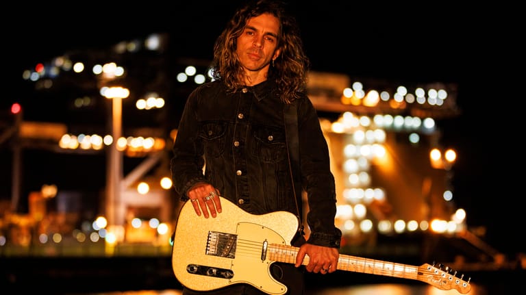 Musician with curly hair holding a cream-coloured electric guitar against a blurred cityscape at night.