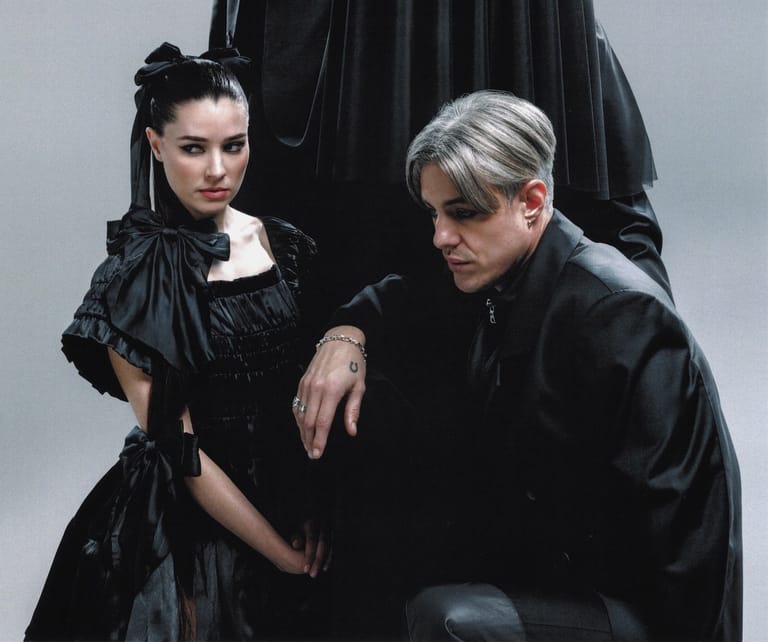 Two performers in dark gothic clothing pose together against a grey backdrop.