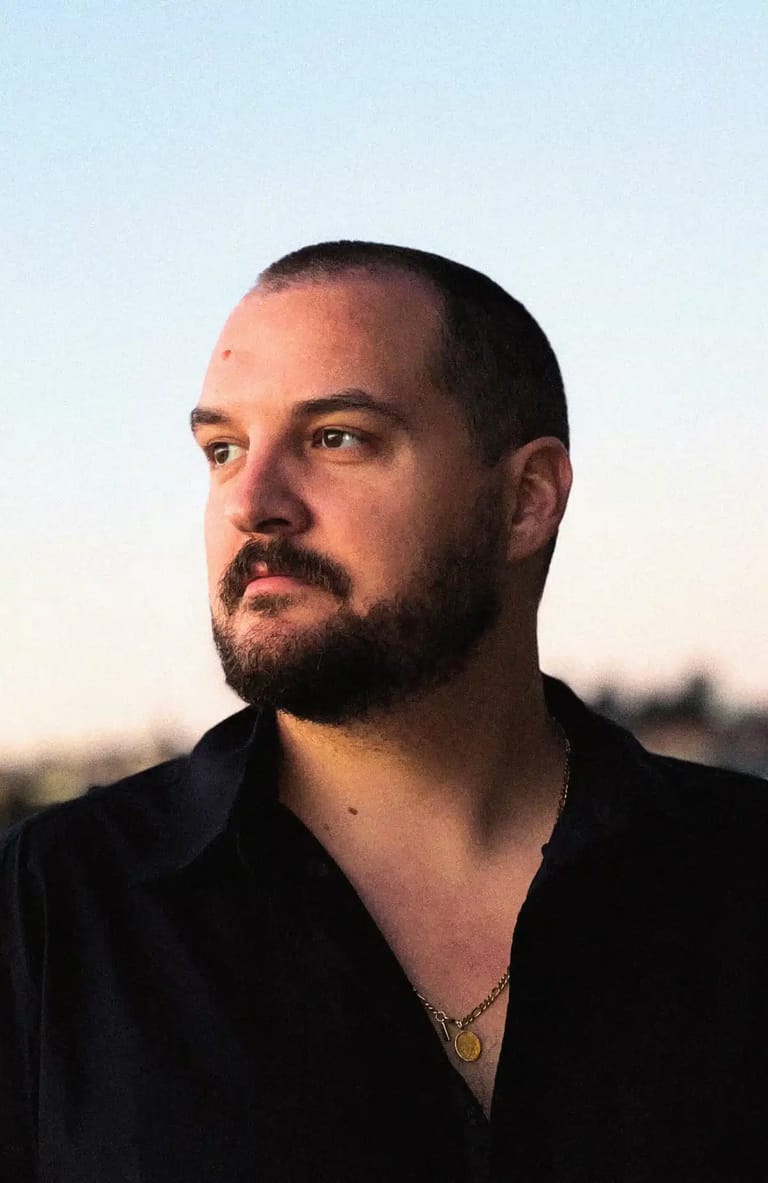 Man with dark hair and beard wearing black shirt, photographed in profile against a blurred sunset background.