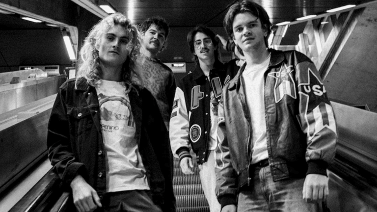 Five band members in leather jackets and rock styling posed in a tour van interior, black and white photograph.