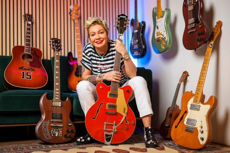 Woman in striped shirt holding red electric guitar surrounded by hanging guitars in music studio.