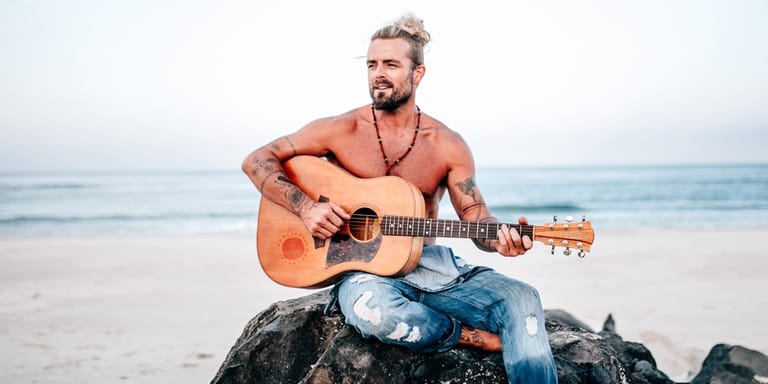 Bearded man with acoustic guitar seated on rocks at a beach, ocean and overcast sky behind him.