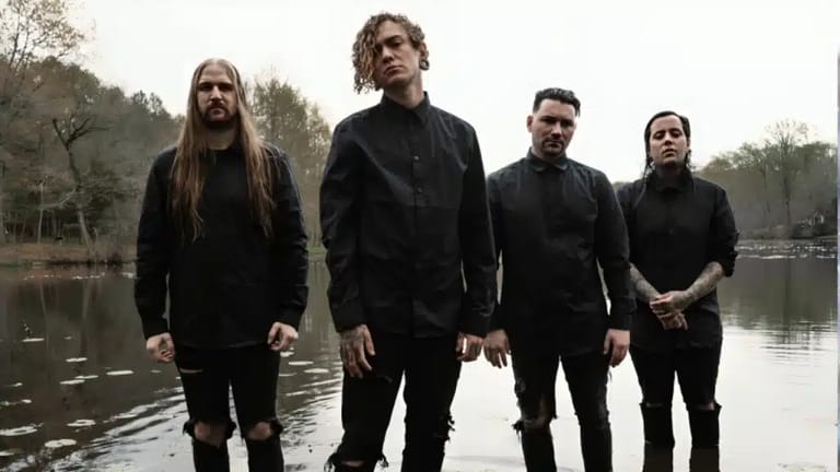Four musicians in black clothing stand together by a still lake surrounded by trees.