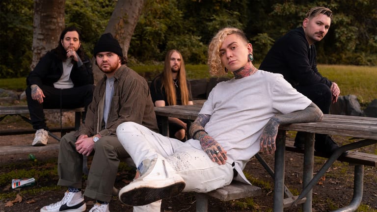 Five musicians in casual attire relaxed on a picnic table at dusk, surrounded by trees.
