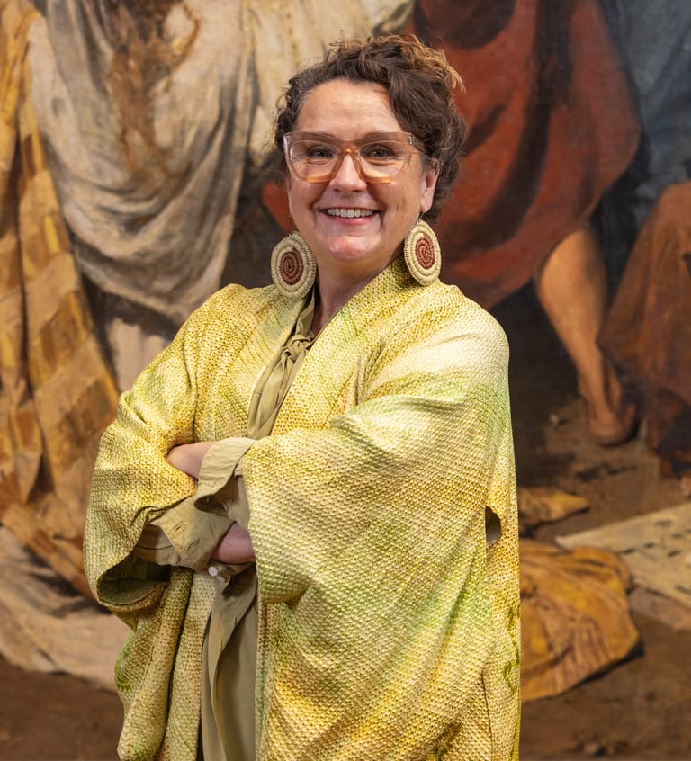 Person wearing yellow textured shirt and large gold hoop earrings smiles in front of draped fabric backdrop.