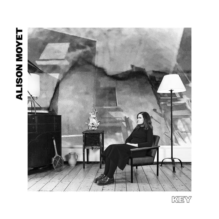 Person seated in chair under dramatic sculptural forms in monochrome studio setting.
