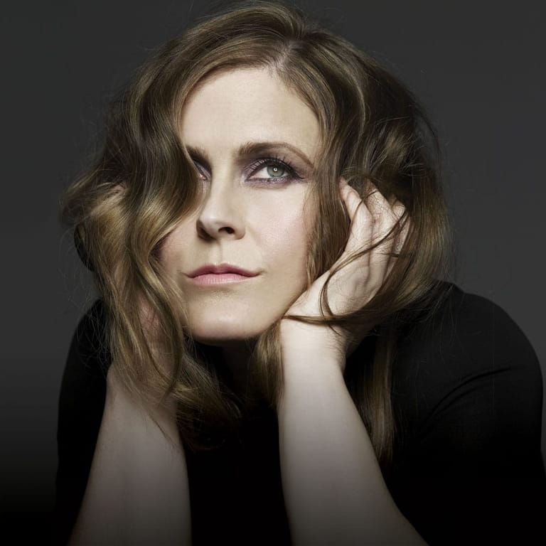 Person with shoulder-length wavy brown hair resting their head on their hand against a dark background.