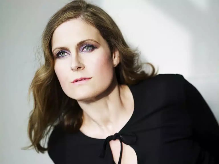 Woman with shoulder-length wavy brown hair and purple eyeshadow wearing a black top against a white background.