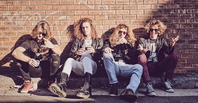 Four musicians sit against a brick wall in casual poses, wearing dark clothing and sunglasses.