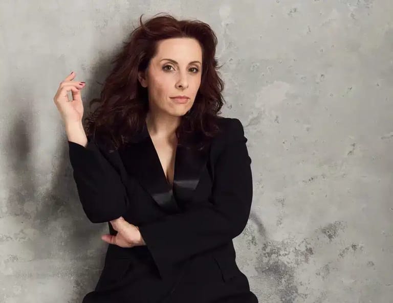 Woman with long dark wavy hair wearing a black blazer against a textured grey wall.